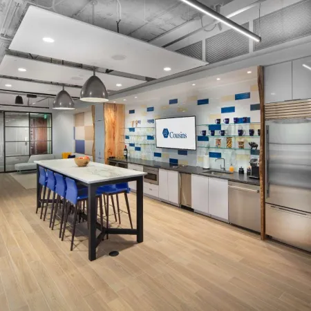 Modern office kitchen with long island, blue chairs, stainless appliances, and patterned backsplash wall.