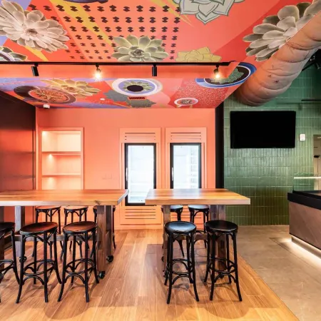 Modern cafe interior with wooden tables, black stools, colorful floral ceiling, green tiled wall, and bright lighting.