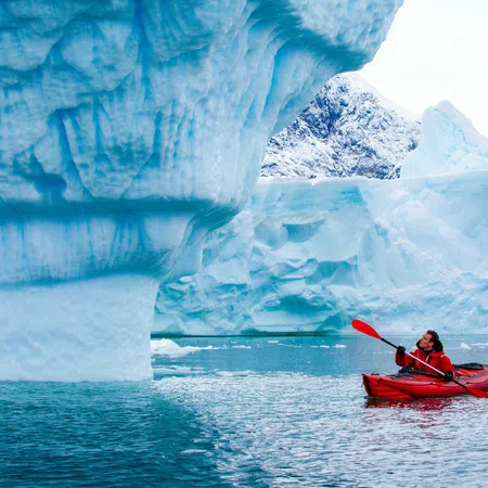 Person in red kayak paddling near large blue icebergs in icy polar waters under a clear sky.