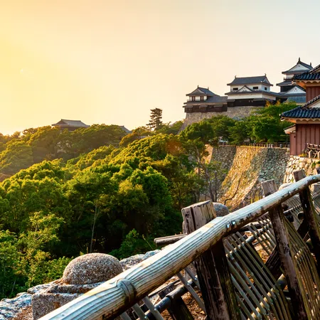Historic Japanese castle on hill with lush green forest and wooden fence pathway at sunset