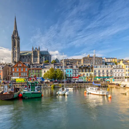Colorful waterfront buildings and boats docked in a calm harbor with a historic cathedral in the background under blue sky.