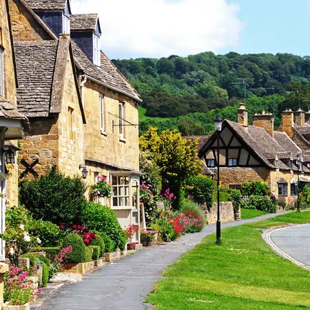 Charming English village street with stone cottages, blooming gardens, and green hills under a blue sky.