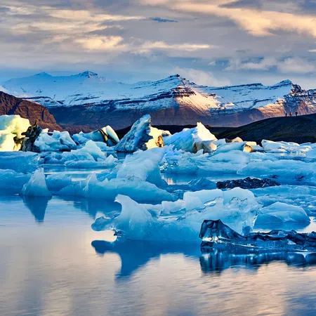 Floating blue icebergs on calm water with snow-capped mountains and cloudy sky at sunset.