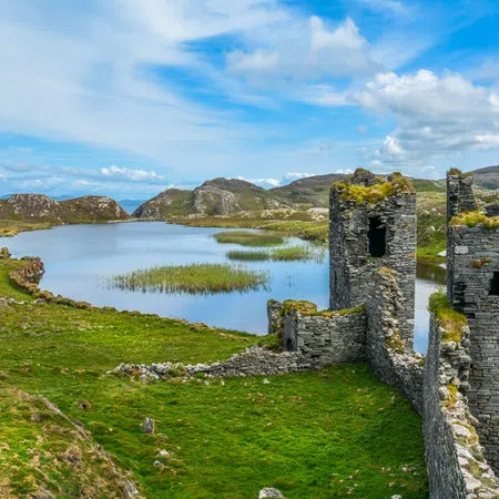 Ancient stone castle ruins beside a calm lake with grassy hills under a partly cloudy sky