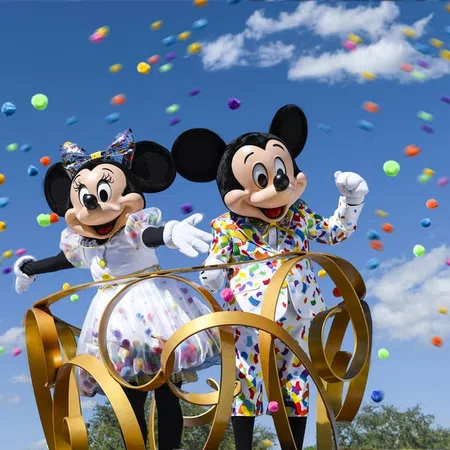 Mickey and Minnie Mouse in colorful outfits surrounded by floating multicolored pom-poms on a bright sunny day.