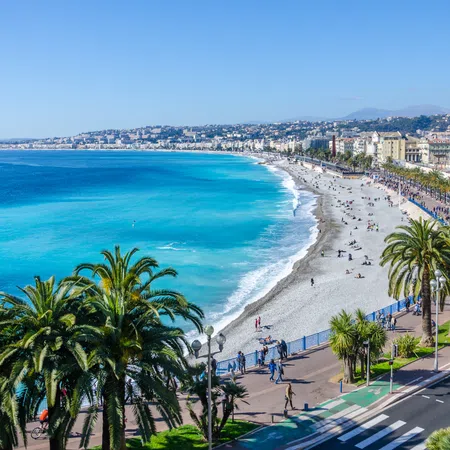 Panoramic view of a Mediterranean beach with turquoise water, palm trees, and a coastal cityscape under clear sky
