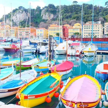 Colorful boats docked in a serene harbor with buildings and green hills in the background under clear skies.