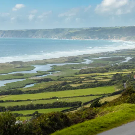 Scenic coastal landscape with winding road, green fields, and blue ocean under cloudy sky