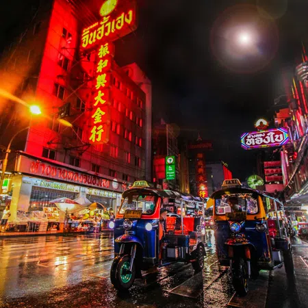 Night street scene with lit tuk-tuks and neon signs in a busy Asian city illuminated by rain-soaked reflections.