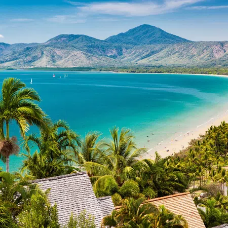 Tropical beach with turquoise water, palm trees, sandy shore, distant mountains, and sailboats under a clear sky