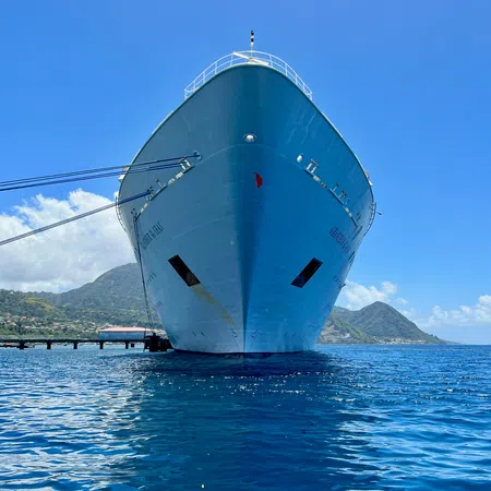 Front view of a large blue cruise ship docked near a tropical island under a clear blue sky.