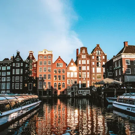 Colorful historic canal houses reflecting in water with sightseeing boats docked under a clear blue sky.