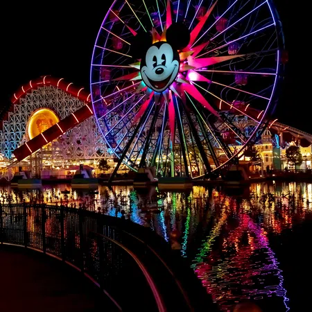 Night view of a brightly lit Mickey Mouse Ferris wheel and roller coaster with colorful reflections on water.