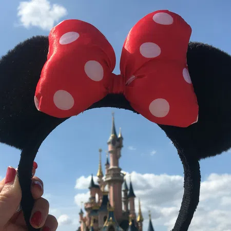 Hand holding Minnie Mouse ears headband with red bow in front of a fairy tale castle under a blue sky.