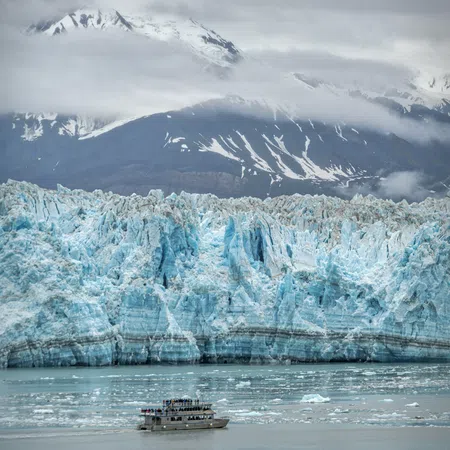 Tour boat near massive blue glacier with snow-capped mountains and clouds in the background