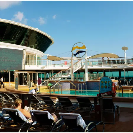 Sunny deck of a Royal Caribbean cruise ship featuring a pool, lounge chairs, and passengers relaxing under blue sky.