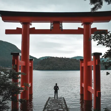 Person standing on a stone pier facing a large red torii gate over a lake with forested hills in the background