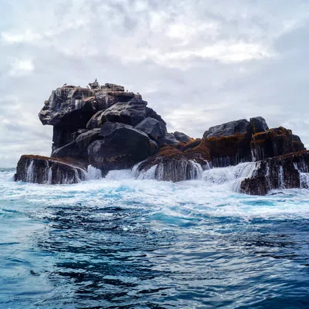 Waves crashing against a rugged rock formation in the ocean under a cloudy sky with blue water.