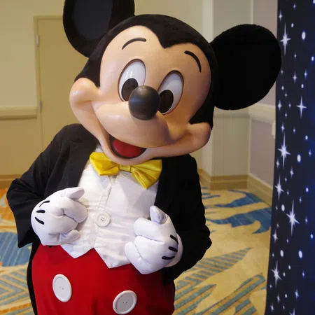 Costumed character of Mickey Mouse in classic red pants, yellow bow tie, and black jacket smiling indoors
