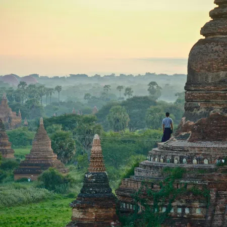 Ancient brick pagodas surrounded by lush greenery and mist with a person standing on a large temple at sunrise.