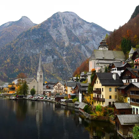 Scenic lakeside village with colorful houses, church steeple, and mountains in autumn foliage under a cloudy sky.