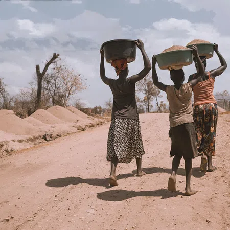 Three women walking barefoot on a dusty road carrying baskets on their heads in a rural area.