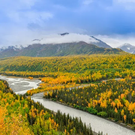 Autumn river winding through forested valley with fall foliage and misty mountains under cloudy sky.