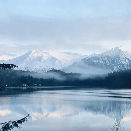 Snow-covered mountains reflected in a calm lake with mist and surrounding pine trees on a cloudy day