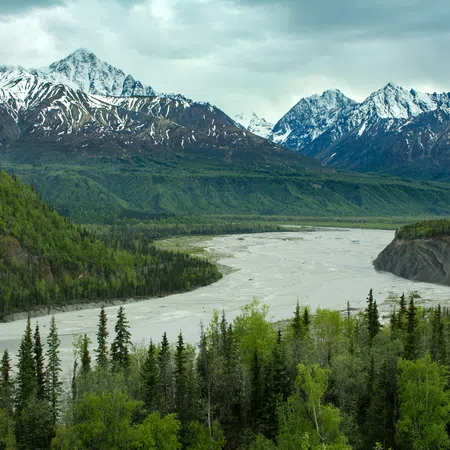 Snow-capped mountains with a winding river and dense green forest under a cloudy sky in a natural landscape.