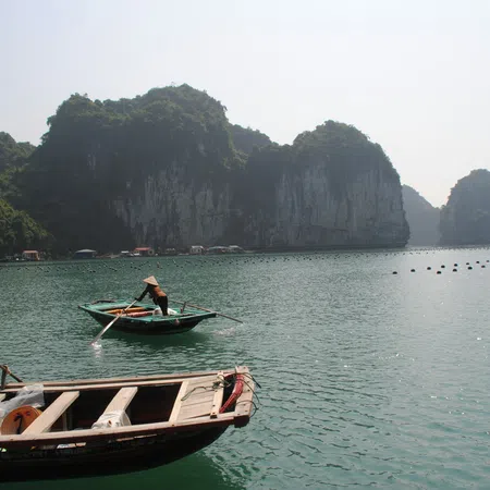 Two people wearing traditional conical hats rowing wooden boats on calm water with limestone karst cliffs in the background.