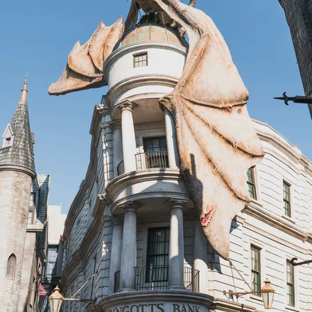 Large dragon perched on Gringotts Bank building with wings spread against clear blue sky.