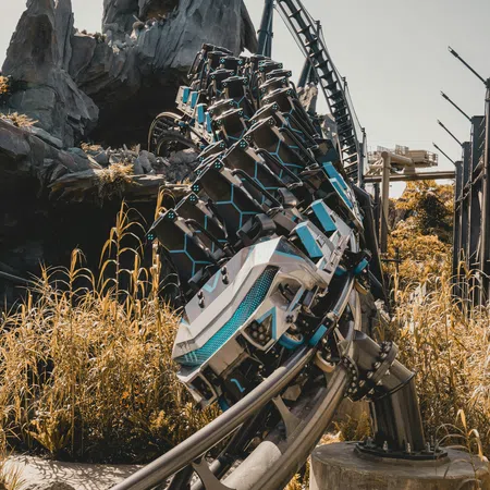 Modern roller coaster car on steel track curving with rocky mountainous backdrop and tall dry grass.