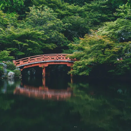 Red wooden bridge over a calm pond surrounded by lush green trees and foliage in a peaceful forest.