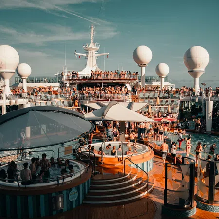 Crowded cruise ship deck with pools, hot tubs, passengers enjoying sunny day and city skyline in background.