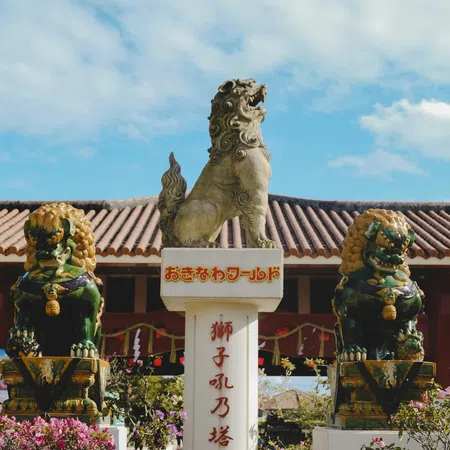 Three Okinawan Shisa statues on pedestals with bright flowers and tiled roof under a blue sky.