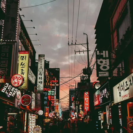 Busy street in a city at dusk with illuminated Korean signs and crowded storefronts under a cloudy sky.