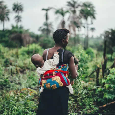 Woman carrying baby in colorful cloth on back walks through lush green farm with palm trees.