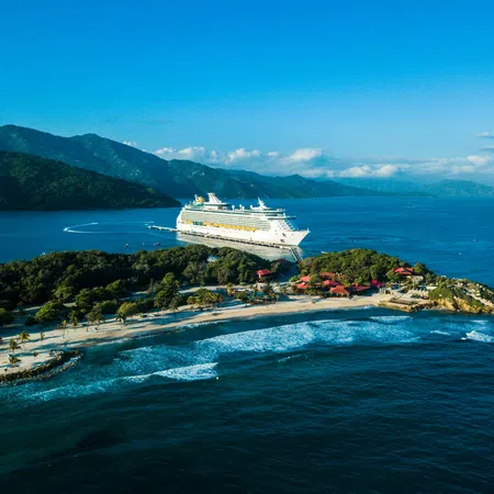 A large cruise ship docked near a lush tropical island with sandy beaches and clear blue ocean waters under a sunny sky.