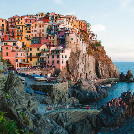 Colorful seaside village perched on rocky cliffs with people enjoying the view and boats in the water below.