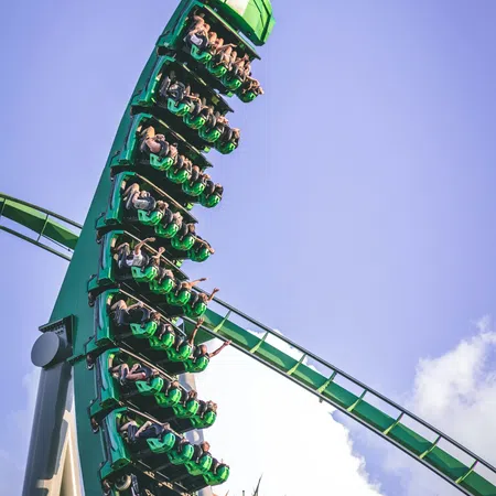 People thrill on a green roller coaster looping against a bright blue sky with amusement park towers below.