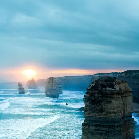 Sunset over the Twelve Apostles limestone stacks along the rugged coastline with waves and cloudy sky.