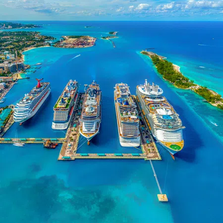 Aerial view of a busy cruise ship port with six large ships docked and turquoise ocean surrounding them