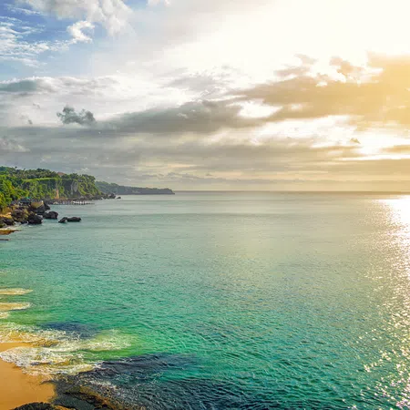 Sunny coastal cliffside with green vegetation, sandy beach, and calm turquoise ocean under a bright sky.