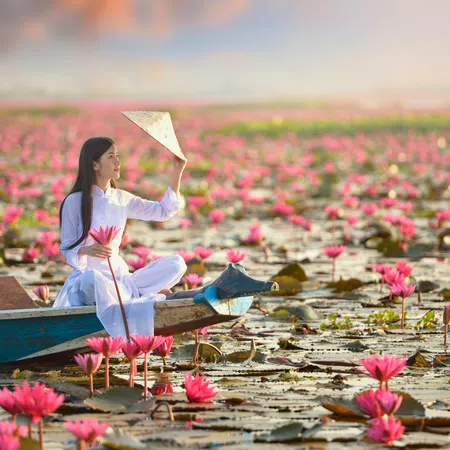 Woman in white áo dài sitting on a boat holding a conical hat among pink lotus flowers on a pond at sunset.