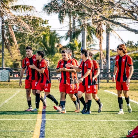 Soccer team in red and black jerseys walking and talking on a sunny field with palm trees in the background.