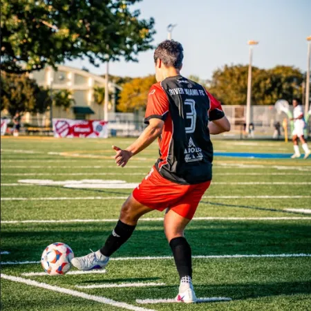 Soccer player in red and black uniform kicking a ball on a sunny outdoor field with trees and buildings in the background.