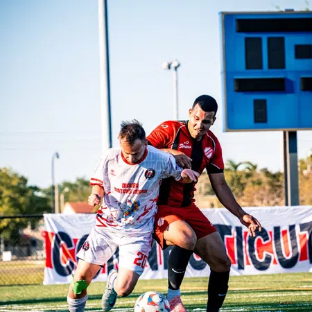 Two soccer players contesting for the ball on a sunny field with a scoreboard and banner in the background