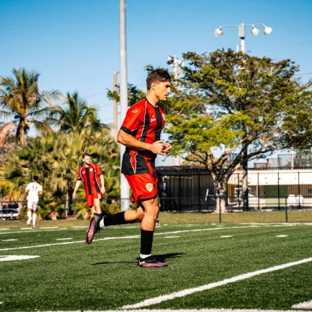 Soccer players in red and white uniforms on a sunny field with palm trees and goalposts in the background.