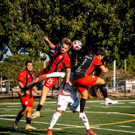 Soccer players in red and white jerseys jump to head the ball on a green field under a tree on a sunny day