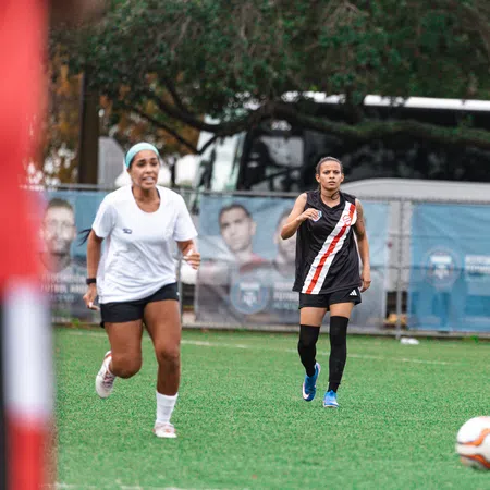 Two female soccer players running on a green field toward a white and orange soccer ball during a game.